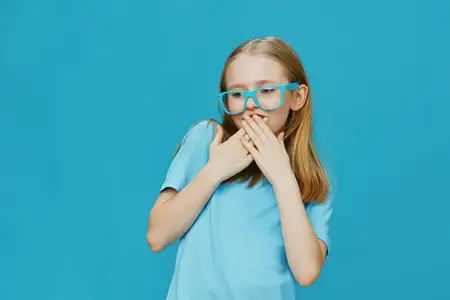 Счастливая юная девушка, демонстрирующая свои длинные волосы и синие очки, игриво закрывает рот на фоне яркого бирюзового фона, отражая радость и невинность