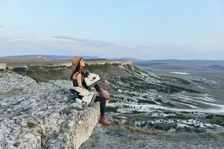 Спокойная женщина играет на гитаре, сидя на скалистом выступе в шляпе, под голубым небом