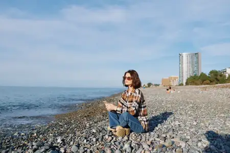 Непринужденный момент на побережье: женщина сидит на каменистом берегу, клетчатая рубашка, солнцезащитные очки, спокойное море и яркое небо, передающие расслабленное одиночество и единение с…