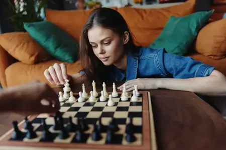Молодая женщина играет в шахматы с мужчиной на диване перед окном в уютной гостиной