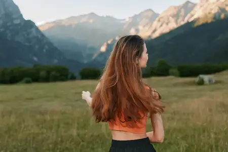 Открытый пейзаж окружает одинокую фигуру в теплой кофте, вглядывающуюся в сторону суровых вершин, с широким небом и безмятежной атмосферой, вызывающей чувство свободы и исследования