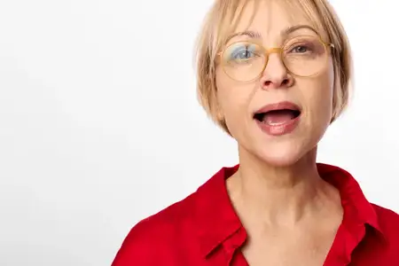 Женщина в очках, портрет крупным планом, удивленное выражение лица, открытый рот, красная блузка, студийное освещение, яркий белый фон, естественные тона кожи, спокойные глаза, современная…
