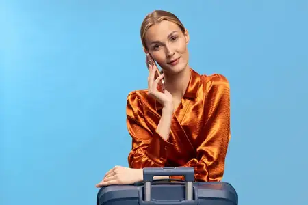 Уверенная молодая женщина в блестящей оранжевой блузке опирается на синий чемодан на фоне голубого задника