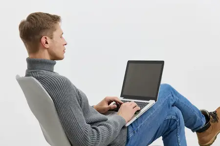 Молодой человек работает за ноутбуком, одетый в серый свитер и синие джинсы, сидит на современном стуле изолированно на белом фоне, сосредоточен на фрилансе, концепция образа жизни цифрового…
