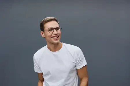 Улыбающийся молодой человек в белой футболке и очках смотрит в сторону со счастливым выражением лица, стоит непринужденно на сером фоне