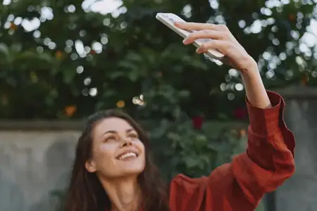 Яркий уличный портрет улыбающейся женщины в красном свитере, делающей селфи со смартфоном среди зелени, передающий расслабленный образ жизни и повседневную радость
