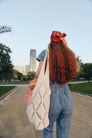 Женщина, ведущая активный образ жизни, одетая в современный бохо-вестерн джинсовый уличный стиль, идёт по дорожке городского парка, неся вязаную сумку-шопер; её рыжие волосы и бандана создают яркое…