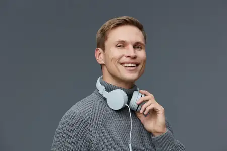 Крупный план радостного мужчины в наушниках, который улыбается, одет в повседневный серый свитер, смотрит в камеру на нейтральном сером фоне