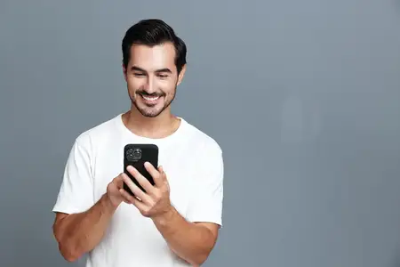 Веселый улыбающийся мужчина увлеченно пользуется смартфоном, на нем простая белая футболка
