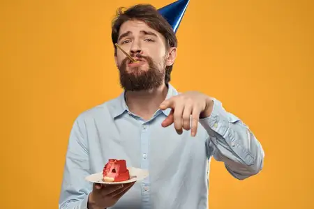 Мужчина с тортом на жёлтом фоне, в праздничном колпаке на день рождения, кадр обрезан