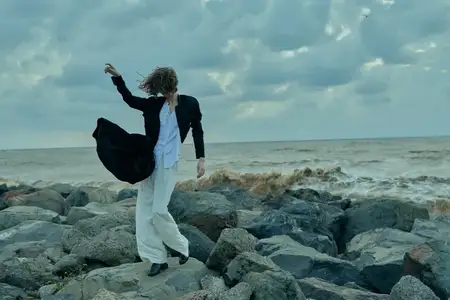 Модное изображение на ветреной береговой линии: развевающийся белый костюм и черное пальто, суровые скалы и облачное небо