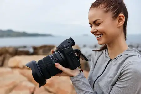 Веселая молодая женщина в серой толстовке занимается фотографией на пляже, воплощая творчество, счастье и любовь к запечатлению моментов