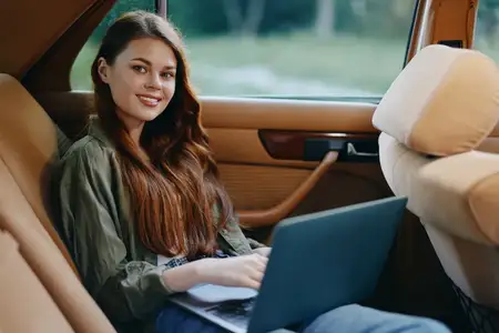 Улыбающаяся молодая женщина с длинными волосами работает за ноутбуком в машине, одетая в повседневную одежду, на фоне спокойного пейзажа, что наводит на мысли о творчестве и удаленной работе