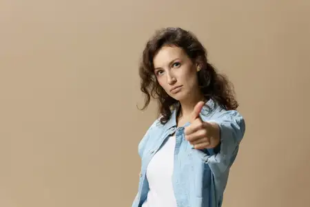 Серьезная уверенная кудрявая красивая женщина в джинсах и повседневной рубашке показывает жест «большой палец вверх» («Нравится») в камеру, позируя изолированно на бежевом пастельном фоне