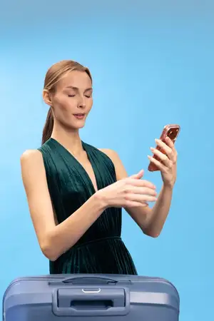 Элегантная молодая женщина в темном платье с чемоданом проверяет смартфон, стоя на голубом фоне