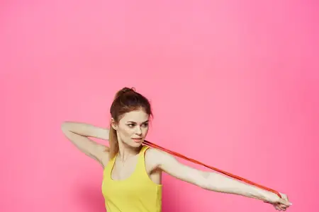 Спортивная женщина прыгает через скакалку, занимаясь упражнениями в жёлтом топе на розовом фоне