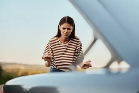 Молодая женщина-водитель в ужасе смотрит на автомобиль, открыв капот