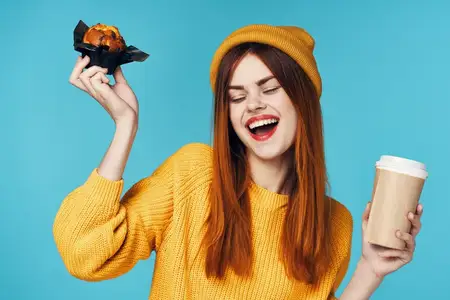 Женщина в желтом свитере и шапке держит в руках чашку кофе и кекс, перекусывая
