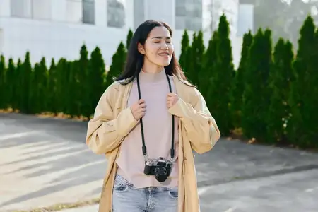 Женщина с фотоаппаратом на улице, улыбается, одета в повседневную одежду: куртку, свитер, джинсы