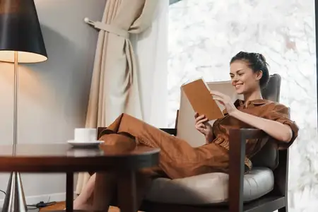 Счастливая молодая женщина в стильном коричневом наряде читает книгу в удобном кресле, потягивая кофе, в окружении безмятежной домашней атмосферы