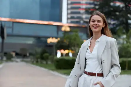 Уверенная женщина в светлом деловом костюме улыбается на открытом воздухе на фоне современных городских зданий и пышной зелени, вызывая ассоциации с успехом и профессионализмом в деловой теме