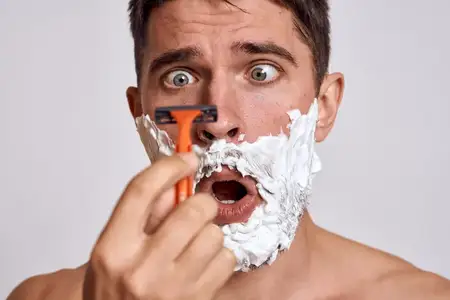 Красивый мужчина с белой пеной для бритья на лице и чистой кожей, с бритвой в руках, занимающийся грумингом