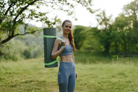 Спортивная женщина в спортивной одежде несет коврик для йоги с радостным выражением лица в безмятежной природной обстановке, что олицетворяет здоровье, фитнес и летнюю жизненную силу