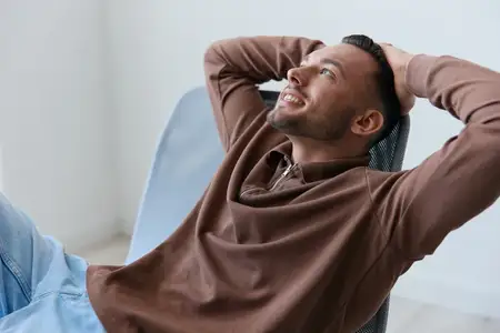 Веселый, довольный, крутой и счастливый молодой человек откидывается на спинку стула, заложив руки за голову, смотрит в сторону, мечтая о будущих каникулах и отдыхе дома