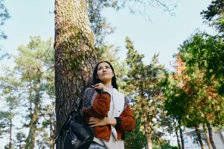 Повседневная уличная сцена: женщина у дерева в светлом парке, наслаждается природой и свежим воздухом, с рюкзаком и теплой курткой, безмятежное лесное настроение и мягкий солнечный свет
