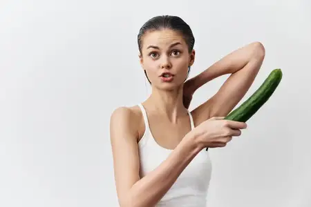 Молодая женщина с удивлённым выражением лица держит в одной руке огурец, а другой касается шеи; она одета в белую майку
