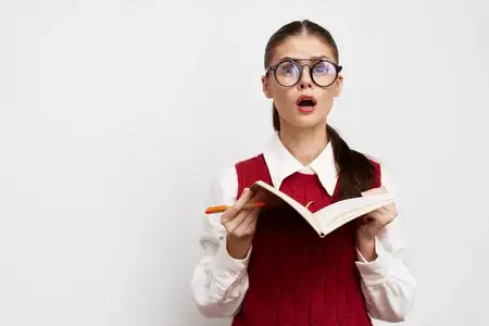 Удивлённая девушка-студентка в очках держит книгу, выглядя потрясённой во время чтения