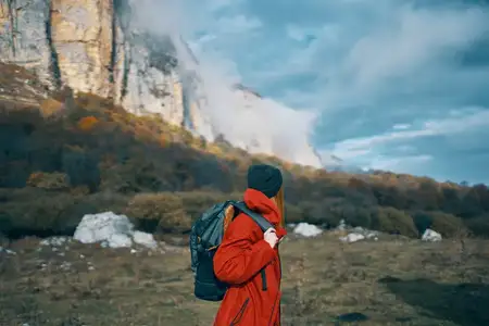 Женщина с рюкзаком и в красной куртке путешествует на фоне голубого неба, высоких гор и скалистого ландшафта