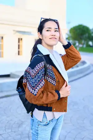 Повседневный уличный стиль: улыбающаяся женщина в теплой вязаной куртке-бомбере, джинсах и с рюкзаком позирует на открытом воздухе у площади