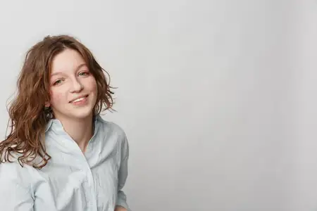 Молодая женщина с вьющимися волосами мягко улыбается в белой рубашке на простом светло-сером фоне