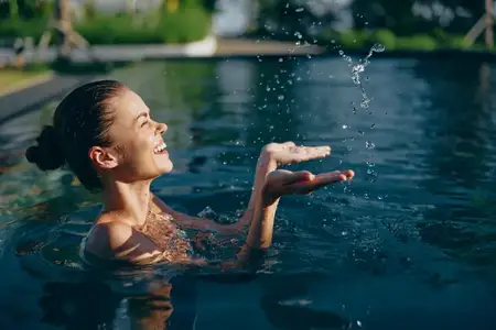 Счастливая женщина в плавательном бассейне играет с брызгами воды, подняв руки вверх, улыбаясь с закрытыми глазами и наслаждаясь летним моментом активного отдыха на свежем воздухе