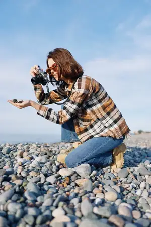 Сцена фотосъёмки на открытом воздухе: женщина-фотограф стоит на коленях среди гладких камней, нацеливая камеру на море под ясным небом