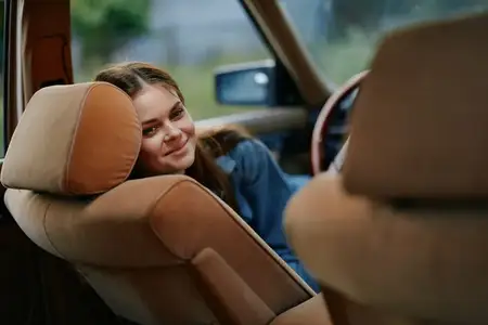Веселая молодая женщина положила голову на спинку сиденья винтажного автомобиля, воплощая беззаботный образ жизни и радостное настроение в безмятежной обстановке на открытом воздухе
