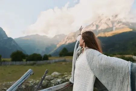 Молодая женщина в уютном белом свитере и серой шали широко раскидывает руки, глядя на живописный горный вид на фоне голубого неба, выражая свободу и связь с природой