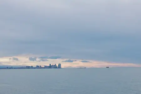 Широкий вид на далекий городской горизонт над спокойным морем, облачный горизонт и мягкий свет, отбрасывающий приглушенное сияние на воду в безмятежной прибрежной панораме