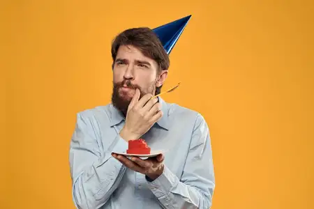 Мужчина держит в руках праздничный торт, на нём колпак, на жёлтом фоне — в день его рождения