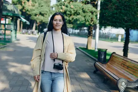 Сцена в парке на открытом воздухе: женщина с фотоаппаратом в легком пальто и джинсах позирует на солнечной дорожке для лайфстайл-фотосессии, представляя естественное выражение лица и расслабленную…