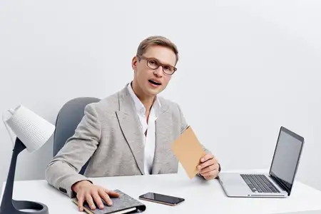 Бизнесмен в очках и официальном костюме работает за столом с ноутбуком, держит папку, выражая удивление или интерес