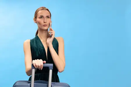 Элегантная женщина в зеленом платье держится за ручку чемодана и разговаривает по мобильному телефону на голубом фоне