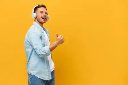 Танцующий счастливый загорелый красивый мужчина в синей базовой футболке и наушниках показывает жест «большой палец вверх» в знак одобрения и позирует изолированно на оранжево-желтом студийном фоне