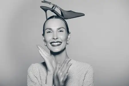 Портрет счастливой светлокожей женщины европейской внешности, улыбающейся с туфелькой на высоком каблуке на голове, в вязаном свитере