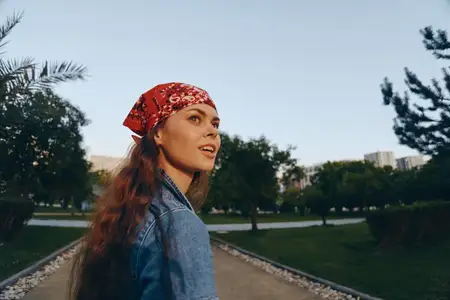 Молодая женщина с красной банданой и длинными волосами в джинсовой куртке идет по зеленому парку среди деревьев