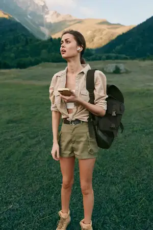 Женщина с рюкзаком в горном лугу держит телефон, на ней шорты и рубашка, в ушах беспроводные наушники