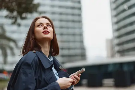 Молодая женщина с длинными каштановыми волосами в тёмном пальто держит в руках смартфон и задумчиво смотрит вверх в городской обстановке, на заднем плане — размытые здания