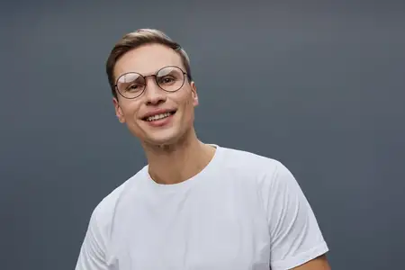 Улыбающийся молодой мужчина европеоидной расы в очках и белой футболке смотрит прямо в камеру с дружелюбным выражением лица, изолирован на темно-сером фоне