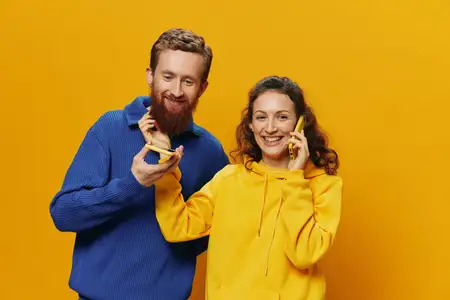 Женщина и мужчина, жизнерадостная пара с телефонами в руках, разговаривают по мобильному телефону, кривая улыбка, веселые, на желтом фоне